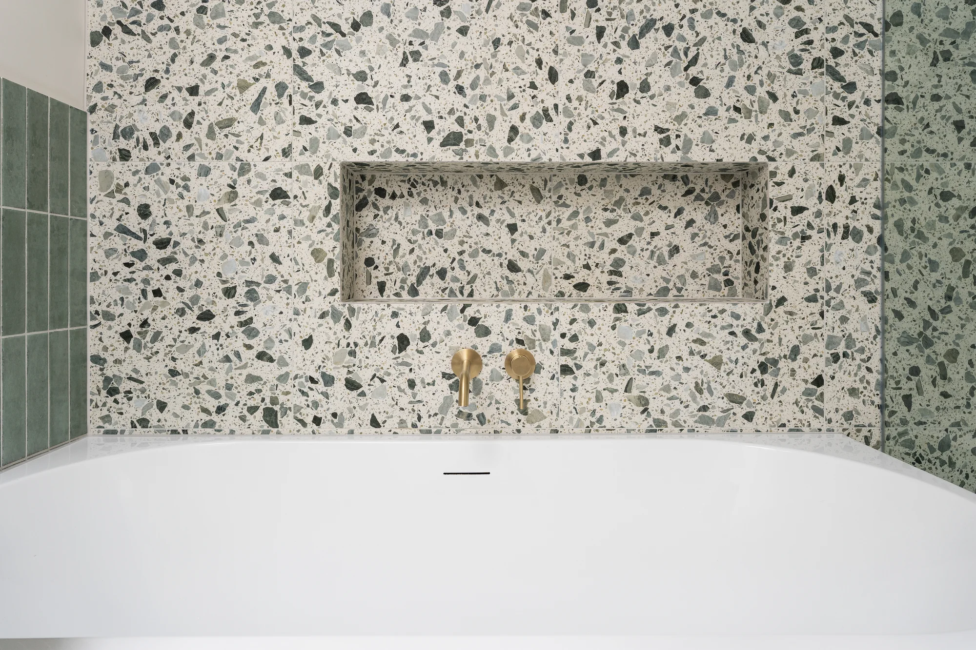 White corner bath beside an emerald tiled wall and recessed shelf